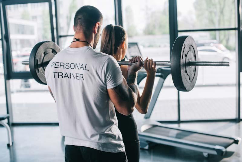 A personal trainer works with a client lifting weights. Learn the pros and cons of starting a personal trainer business at EliteTrainr.com.